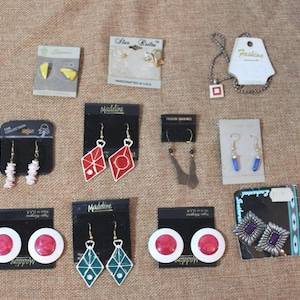 May include: A selection of vintage earrings and a necklace. The earrings showcase diverse designs, including geometric shapes, floral details, and dangling beads. The necklace has a square pendant. The items are arranged on a textured, neutral-toned surface.