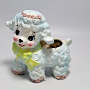 Könnte beinhalten: Vintage Keramik-Lamm-Pflanzgefäß, hellblau mit lockiger Textur. Es hat eine gelbe Schleife, rosa Akzente und einen Blumenkranz. Die Rückseite ist ausgehöhlt, um kleine Pflanzen oder Gegenstände aufzunehmen.