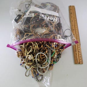 Peut inclure: Un sac en plastique transparent rempli de nombreux colliers de perles de différentes couleurs, dont le marron, le beige et le bleu. Une règle en bois est positionnée à droite du sac. Le sac est fermé par une fermeture éclair rose.