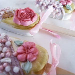 Pu&ograve; includere: Un set di quattro cake pops decorati con glassa rosa e bianca, codette e fiori. I cake pops sono su stecchi e hanno nastri rosa legati intorno.