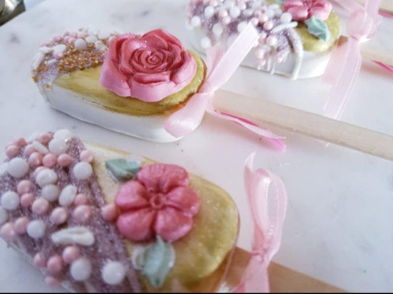 Pu&ograve; includere: Un set di quattro cake pops decorati con glassa rosa e bianca, codette e fiori. I cake pops sono su stecchi e hanno nastri rosa legati intorno.