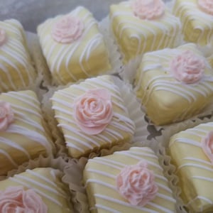 Op de afbeelding: Een close-up van een groep kleine, vierkante taarten versierd met witte glazuur en roze rozen. De taarten zijn gerangschikt op een wit oppervlak.