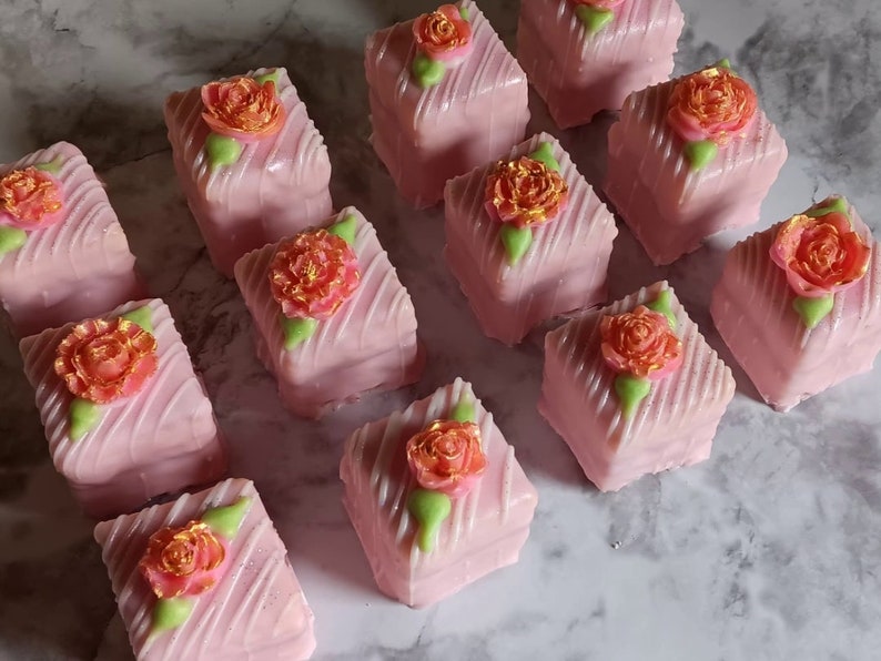 Op de afbeelding: Een verzameling roze en witte petit fours met gouden en groene bloemdecoraties.
