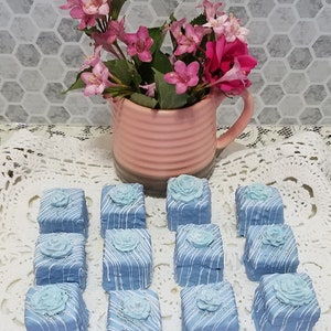 Op de afbeelding: Twaalf blauwe en witte geglazuurde petit fours met een delicaat wit rozenontwerp erop, gerangschikt op een witte kantonderzetter. Een roze en witte bloemstuk staat op de achtergrond.