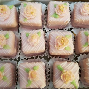 Op de afbeelding: Een doos met roze en witte petit fours met gouden accenten. Elke petit four is versierd met een roze roos en groene bladeren.