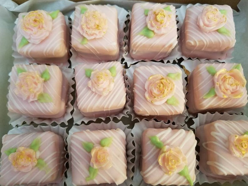 Op de afbeelding: Een doos met roze en witte petit fours met gouden accenten. Elke petit four is versierd met een roze roos en groene bladeren.