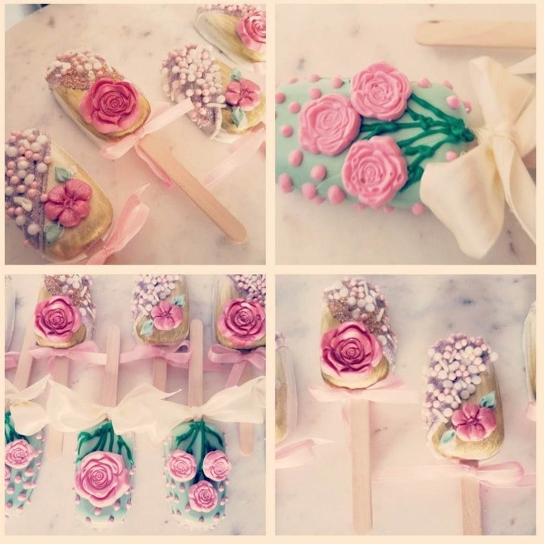 Pu&ograve; includere: Una collezione di cake pops decorati con fiori rosa e bianchi. I cake pops sono su bastoncini e hanno una base dorata. Alcuni dei cake pops sono decorati con un motivo floreale verde e bianco.