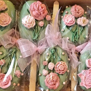 Pu&ograve; includere: Sei cake pops verdi decorati con fiori rosa e foglie verdi, legati con un nastro rosa e avvolti in plastica trasparente.