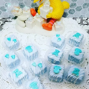 Op de afbeelding: Blauwe en witte geglazuurde cake pops versierd met babyvoetjes, rompertjes en teddyberen. De cake pops zijn gerangschikt op een witte kantonderzetter.