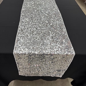 May include: A silver sequin table runner displayed on a black tablecloth. The runner is rectangular and covered in small, reflective sequins. The sequins create a shimmering effect, ideal for weddings or special events.
