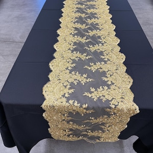 Puede incluir: Un camino de mesa de encaje dorado con un diseño floral se muestra sobre un mantel negro. El camino de mesa tiene un borde festoneado y está centrado en la mesa. El encaje es intrincadamente detallado y añade un toque de elegancia.