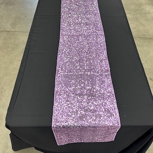 Könnte beinhalten: Ein Tischläufer aus lavendelfarbenen Pailletten mit einem dezenten Schimmer. Der Läufer liegt auf einer schwarzen Tischdecke.