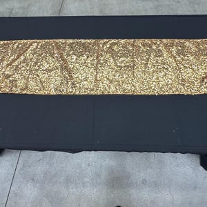 Può includere: Un runner da tavolo nero con una striscia di paillettes dorate al centro.