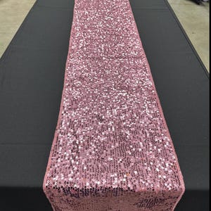 Puede incluir: Un camino de mesa de lentejuelas rosa. El camino de mesa está hecho de pequeñas lentejuelas iridiscentes que crean un efecto brillante.