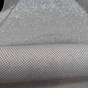 May include: A roll of shimmering silver fabric with a net-like pattern. The fabric is densely covered in small, sparkling embellishments, creating a dazzling, reflective surface. The material appears to be lightweight and suitable for various craft and design projects.