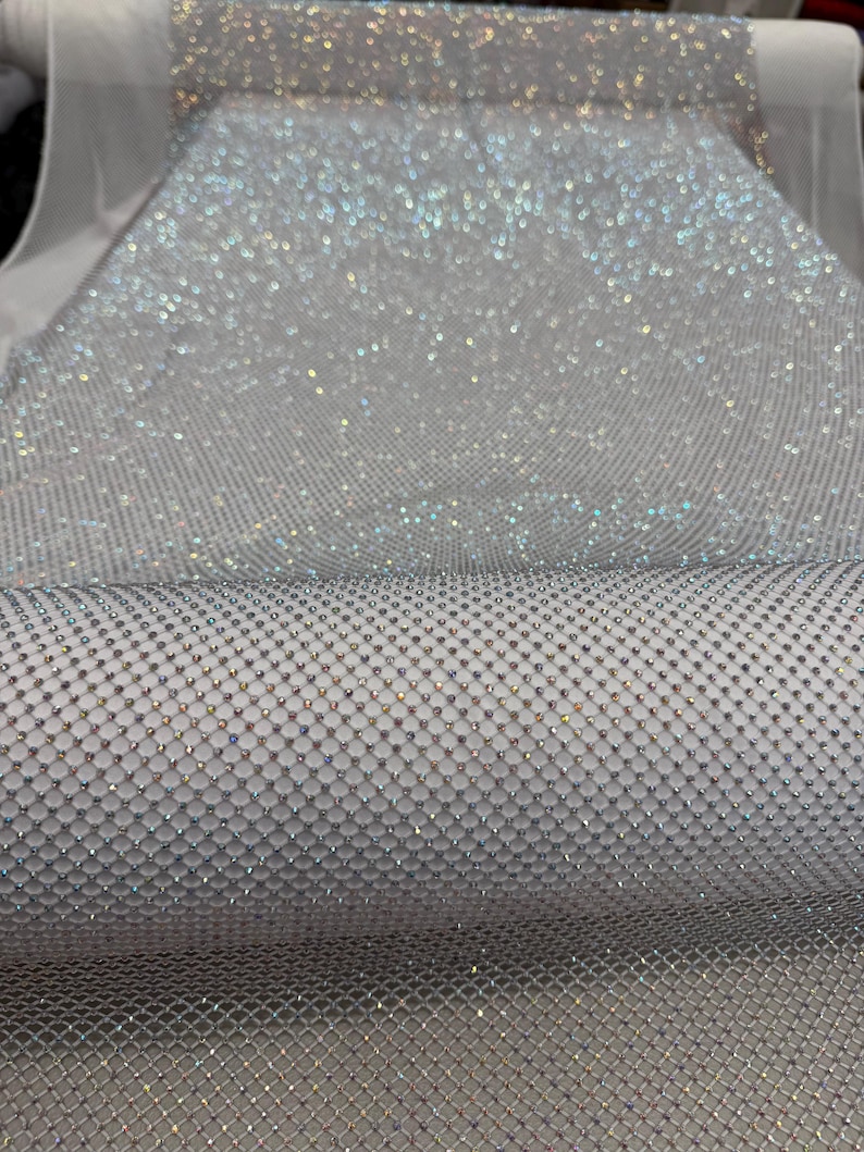 May include: A roll of shimmering silver fabric with a net-like pattern. The fabric is densely covered in small, sparkling embellishments, creating a dazzling, reflective surface. The material appears to be lightweight and suitable for various craft and design projects.