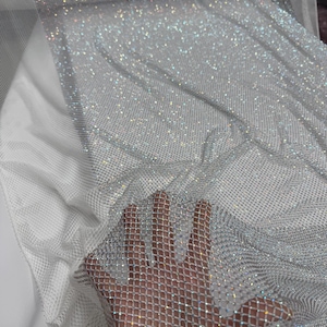 May include: A shimmering silver mesh fabric with a diamond pattern. The fabric is covered in small, sparkling embellishments, creating a glittery effect. The material is draped over a white surface, showcasing its texture and light-reflecting qualities.