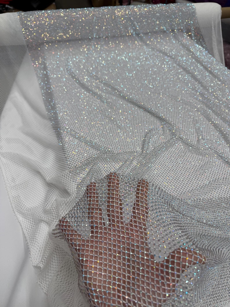 May include: A shimmering silver mesh fabric with a diamond pattern. The fabric is covered in small, sparkling embellishments, creating a glittery effect. The material is draped over a white surface, showcasing its texture and light-reflecting qualities.