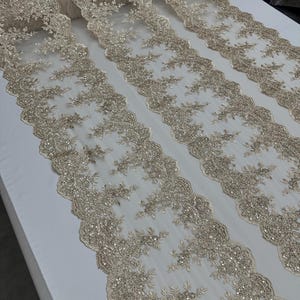 May include: Beige embroidered lace fabric with a scalloped edge and silver sequins. The fabric is rolled up on a white surface.