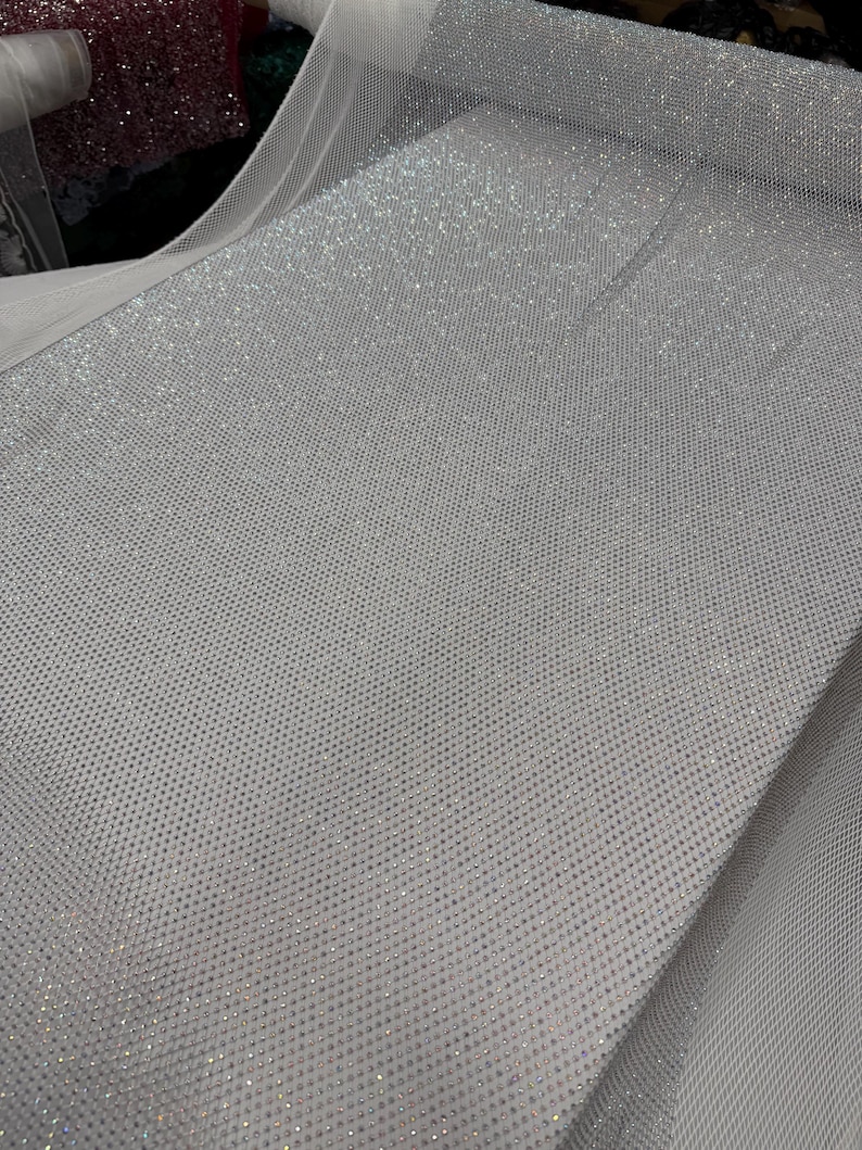 May include: A roll of white fabric with a dense pattern of small, sparkling embellishments. The fabric has a fine mesh backing, and the surface is covered in tiny, reflective elements, creating a shimmering effect. The fabric is suitable for apparel and craft projects.
