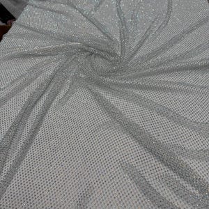 May include: A close-up of a silver fabric with a net-like pattern. The fabric is covered in small, sparkling embellishments, creating a shimmering effect. The material is draped, showing folds and texture. The overall appearance is elegant and decorative.