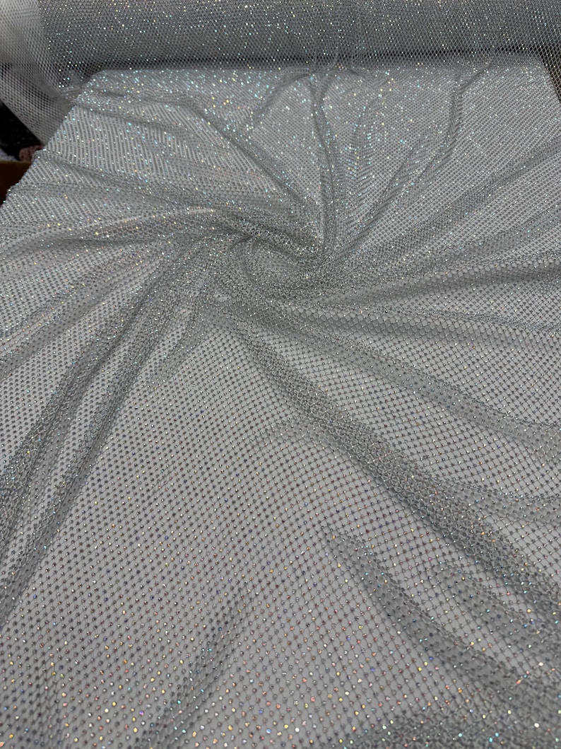 May include: A close-up of a silver fabric with a net-like pattern. The fabric is covered in small, sparkling embellishments, creating a shimmering effect. The material is draped, showing folds and texture. The overall appearance is elegant and decorative.