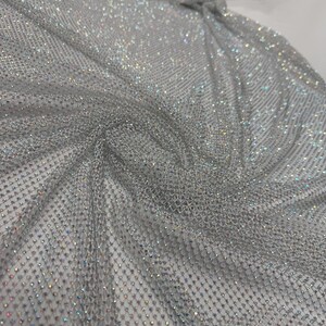 May include: A shimmering silver fabric with a net-like structure. The fabric is covered in small, sparkling embellishments that catch the light, creating a dazzling effect. The material is draped, showing its flexibility and potential for use in apparel or decorative projects.