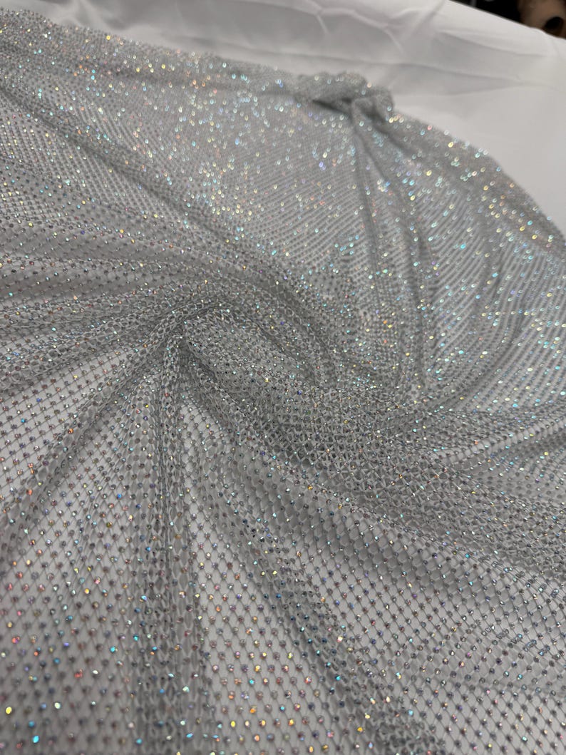 May include: A shimmering silver fabric with a net-like structure. The fabric is covered in small, sparkling embellishments that catch the light, creating a dazzling effect. The material is draped, showing its flexibility and potential for use in apparel or decorative projects.