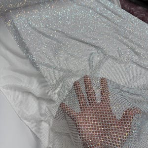 Op de afbeelding: Een close-up van een glinsterende, zilverkleurige stof met een netachtige structuur. De stof is bedekt met kleine, reflecterende versieringen, waardoor een sprankelend effect ontstaat. De stof is gedrapeerd over een wit mesh materiaal.