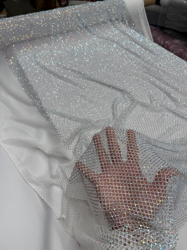 May include: A close-up of a shimmering, silver fabric with a net-like structure. The fabric is covered in small, reflective embellishments, creating a sparkling effect. The fabric is draped over a white mesh material.