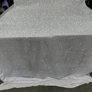 May include: White fabric with small, white sequins scattered across the surface. The fabric is folded and draped over a surface.