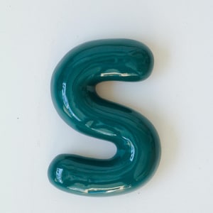 May include: A teal green letter S made of ceramic. The letter has a glossy finish and a rounded, bubbly shape.