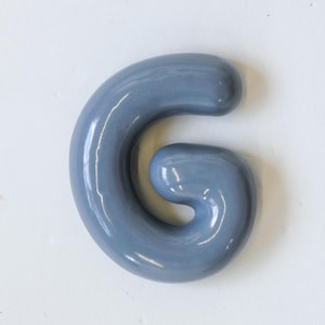 May include: A light blue ceramic letter G. The letter is sculpted in a rounded, organic style.