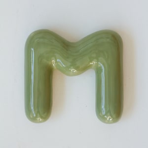 May include: A green ceramic letter M. The letter is glossy and has a rounded, bubbly shape.