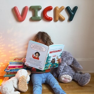 May include: A child reads a book titled "NUTSHIMIT" on a wooden floor, surrounded by books and soft toys. Above, the name "VICKY" is displayed in colourful, 3D letters on a white wall, creating a playful atmosphere.