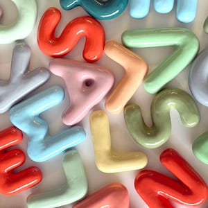 May include: A selection of brightly coloured ceramic letters in pastel hues, such as red, blue, green, and yellow. The letters are scattered, with the word "CHAMPAGNE" printed in black at the bottom.