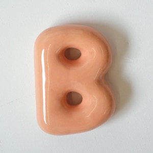 May include: A peach-colored, glossy ceramic letter "B" against a white background. The letter has a rounded, bubble-like design with two circular holes. The smooth surface reflects light, highlighting its curves. This decorative piece could be used for home decor or craft projects.