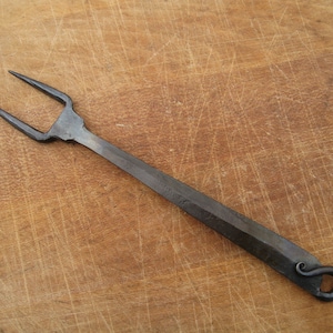 May include: A black metal fork with a long handle and a loop at the end. The fork has three tines and is handcrafted.