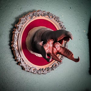May include: A wall-mounted sculpture featuring a dark, textured creature head with an open, multi-jawed mouth protruding from a circular, ornate silver-colored frame. The background inside the frame is a deep red velvet-like material.