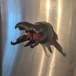 May include: A dark green, creature-like magnet with a gaping mouth and red interior, attached to a metallic surface. The magnet's form casts a shadow, enhancing its three-dimensional appearance. The design is reminiscent of a sea monster.