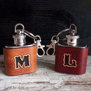 Op de afbeelding: Twee leren hipflasks met zilveren doppen en sleutelhangers. De fles aan de linkerkant is bruin met de letter "M" erop gegraveerd. De fles aan de rechterkant is bordeauxrood met de letter "L" erop gegraveerd.