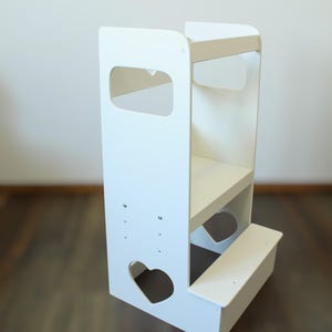Può includere: Sgabello a gradini in legno bianco con due gradini e un'apertura a forma di cuore sul davanti. Il gradino superiore ha uno scaffale e una barra per appendere oggetti.