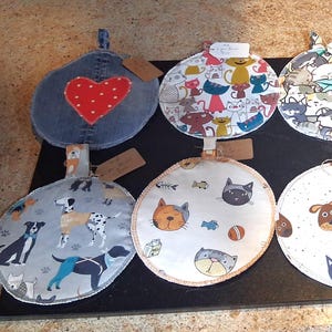 May include: Ten circular fabric pot holders arranged in two rows. Designs include denim with a red heart, various cat and dog patterns, a geometric quilt print, and a blue denim circle featuring a small pocket with a red polka dot fabric accent.