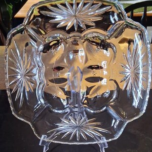 May include: Clear glass serving dish with starburst and floral etched designs. The dish is square with scalloped edges and is displayed on a clear plastic stand. A decorative piece for serving food or displaying items.
