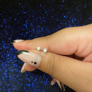 May include: A pair of small, round, clear gemstone stud earrings. The earrings are held between two fingers against a dark blue, glittery background. The gemstones are set in a silver or white metal.
