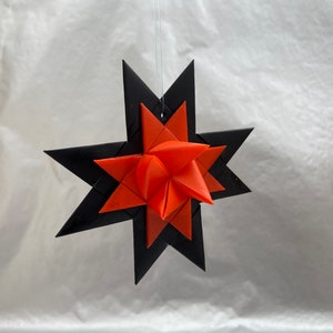 May include: A hanging star-shaped ornament with a layered design. The outer layer is black, and the inner layer is orange. The ornament is suspended by a thin, nearly invisible string, suggesting it's for decoration, possibly for the holidays.