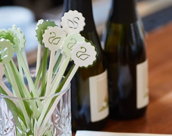 May include: A glass jar filled with green and white stir sticks with a swan design. The stir sticks are arranged in a fan shape. Four bottles of sparkling wine are in the background. The bottles have black caps and are partially obscured by the jar. A white card with the text "You're a limited edition" is in the foreground.