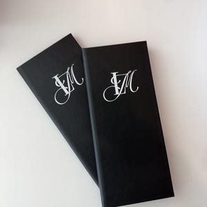May include: Two rectangular black menu covers featuring a white monogram with the letters L and M in a stylized script font. The covers have a smooth, matte texture and are positioned at an angle, overlapping each other on a plain white surface.