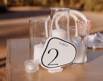 May include: A white, organic-shaped table number sign with a black border and the text "TABLE 2" in black. It is displayed alongside three clear glass cylindrical candle holders containing white pillar candles and a small ribbed glass votive.