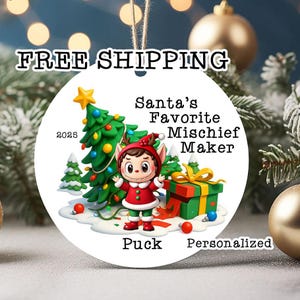May include: A round white Christmas ornament featuring a cartoon elf in a red and green outfit standing next to a decorated Christmas tree and a green gift box. The text reads Santa's Favorite Mischief Maker, 2025, and the name Puck.